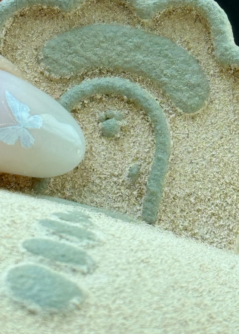 A close-up of a hand pressing a textured, beige silicone or rubber stamp into fine sand, leaving a pale greenish-gray imprint.