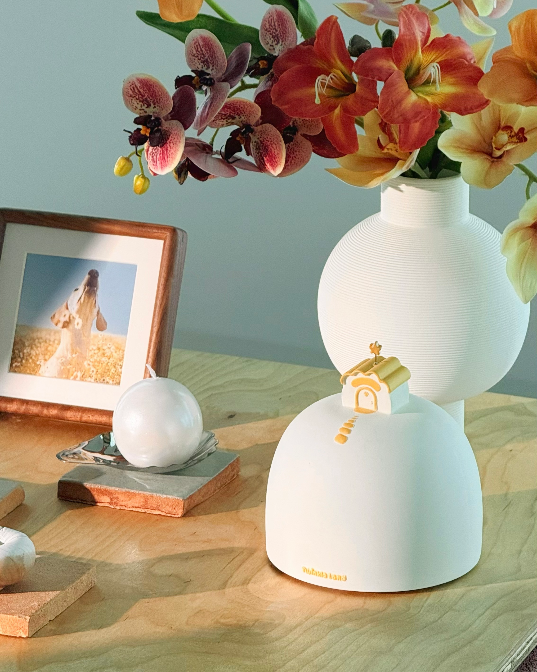 A white ceramic pet urn with a small house detail on the lower tier, filled with orange, pink, and yellow flowers, set on a wooden table next to a framed picture.