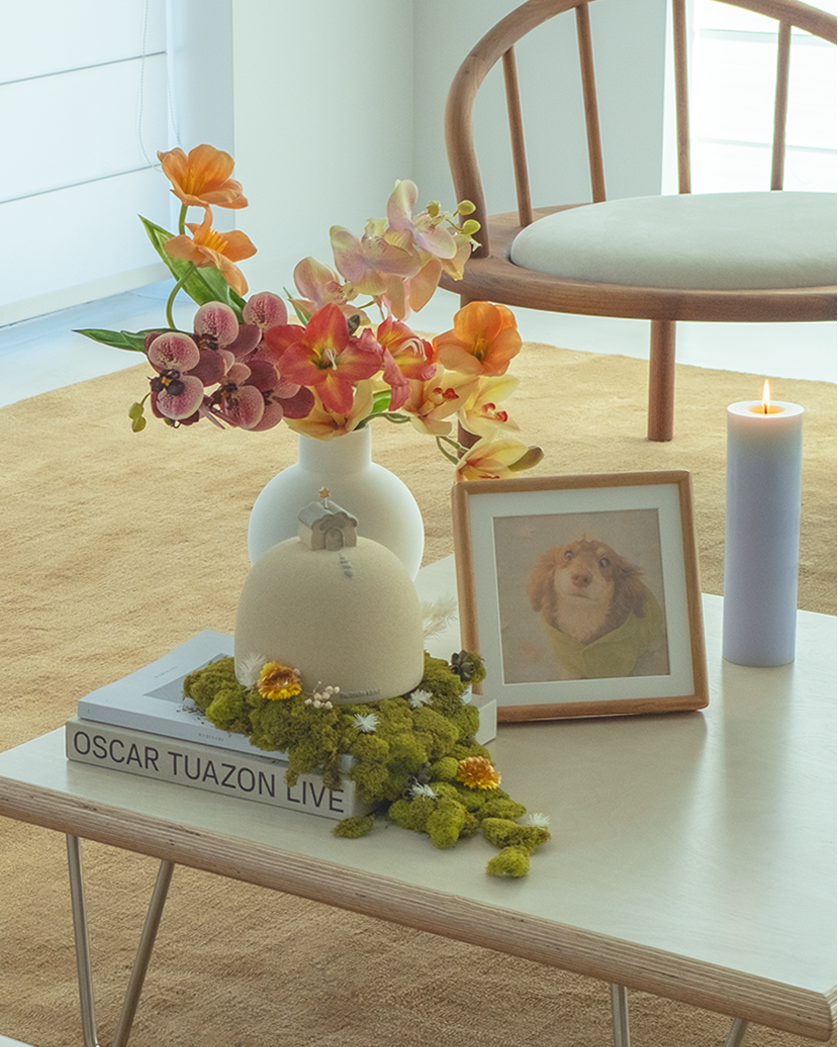 Small table with flowers, a pet urn, and a framed picture in a room with a chair and window.